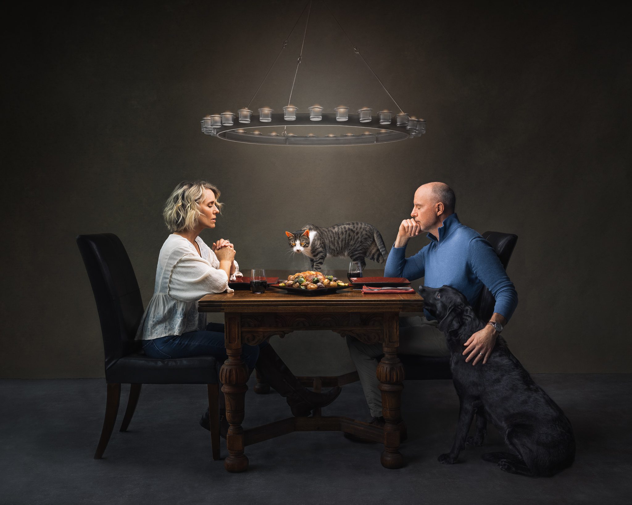 Couple praying at dinner table with cat on table and dog faithfully at dad's side